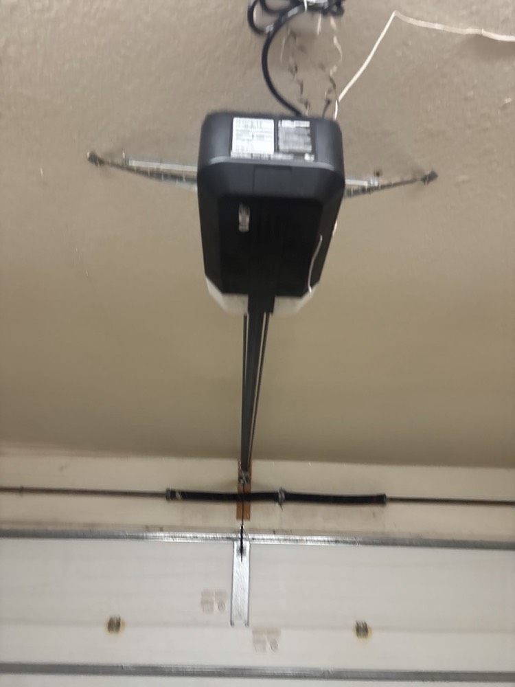 Black garage door opener motor mounted to garage ceiling with connecting wires and hardware.
