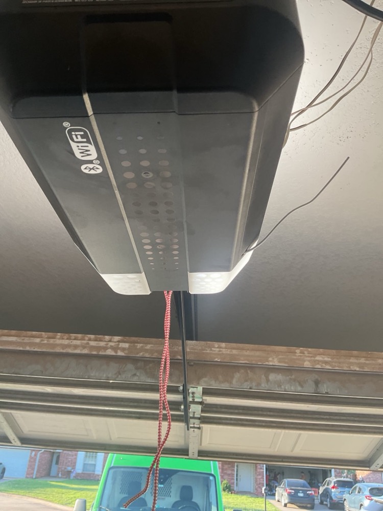 Black WiFi-enabled garage door opener mounted on garage ceiling with wiring visible.