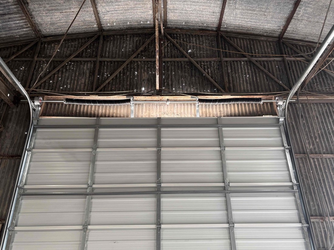 Gray sectional garage door installed in industrial garage with metal roof framework visible above.