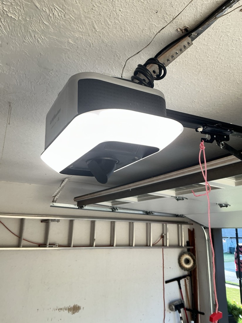 Installed garage door opener with white LED light panel and red safety sensor cord